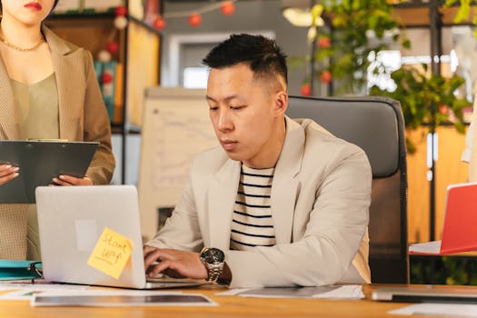 Asian man focusing on work in a modern office, engaged with a laptop, symbolizing a busy start-up environment.