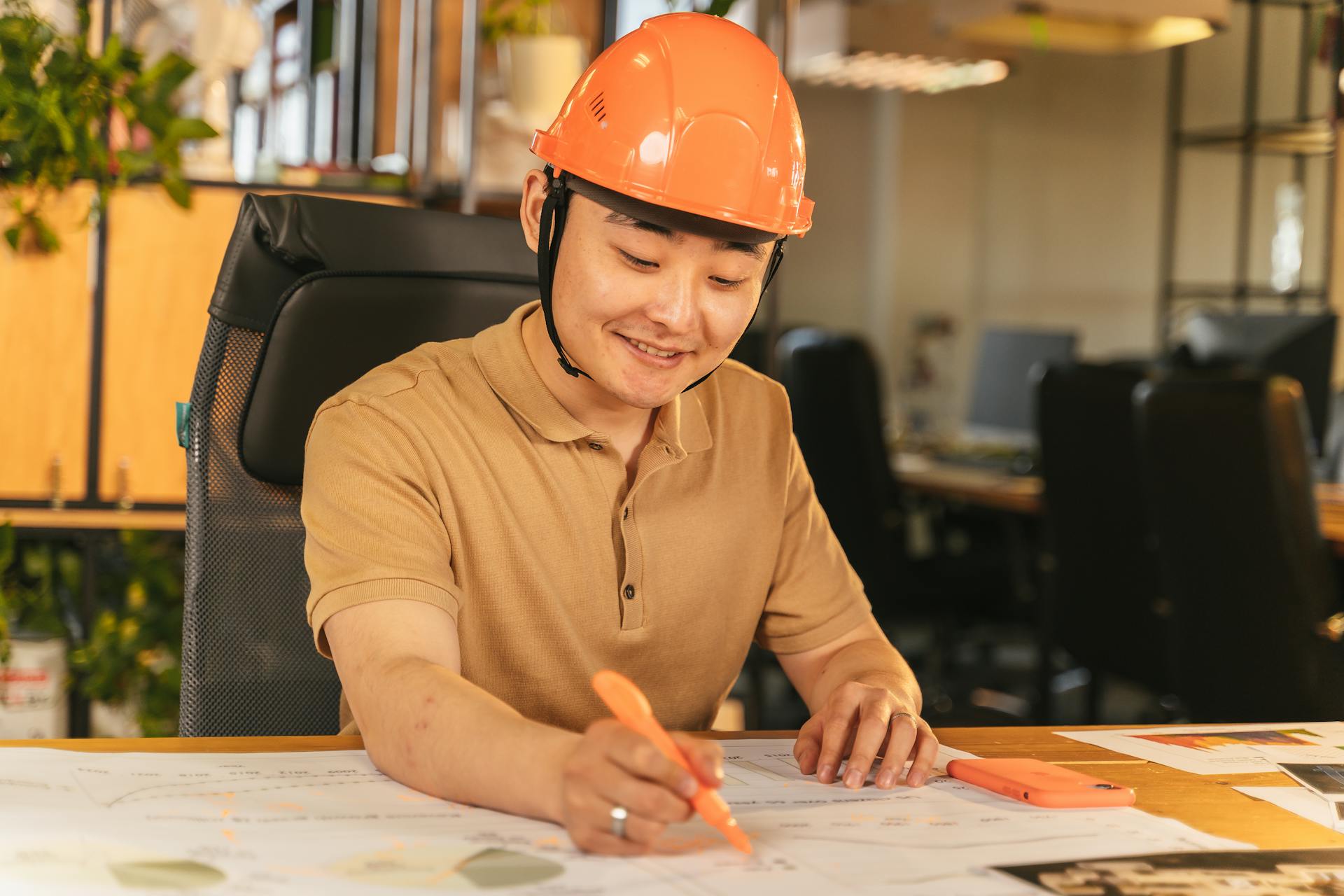 Smiling engineer in hard hat studies blueprints at an office desk, highlighting construction and planning.