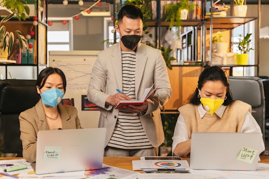 Colleagues wearing masks working together in an office setting with laptops and documents.