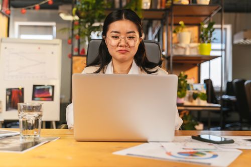 Free Asian woman with eyeglasses working on a laptop in a modern office setting, surrounded by charts and plants. Stock Photo