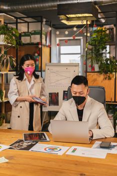 Two professionals in masks collaborate at office desk with charts and laptops.