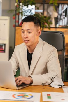 Asian man in white coat typing on a laptop at a wooden desk, focused on work.