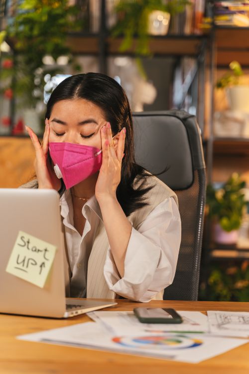 Free Woman with mask feeling tired at her desk in an office setting. Stock Photo