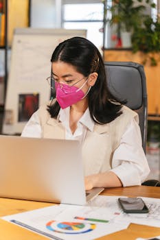 Professional woman wearing a face mask working on her laptop in an office setting.