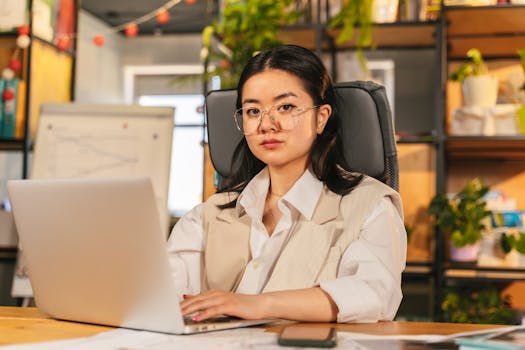 Confident businesswoman with glasses focusing on work using a laptop in a modern office setting.