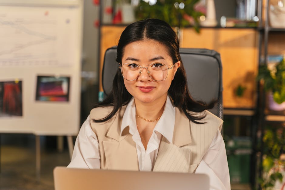 Portrait of a woman in an office setting, confidently using a laptop with charts in the background.