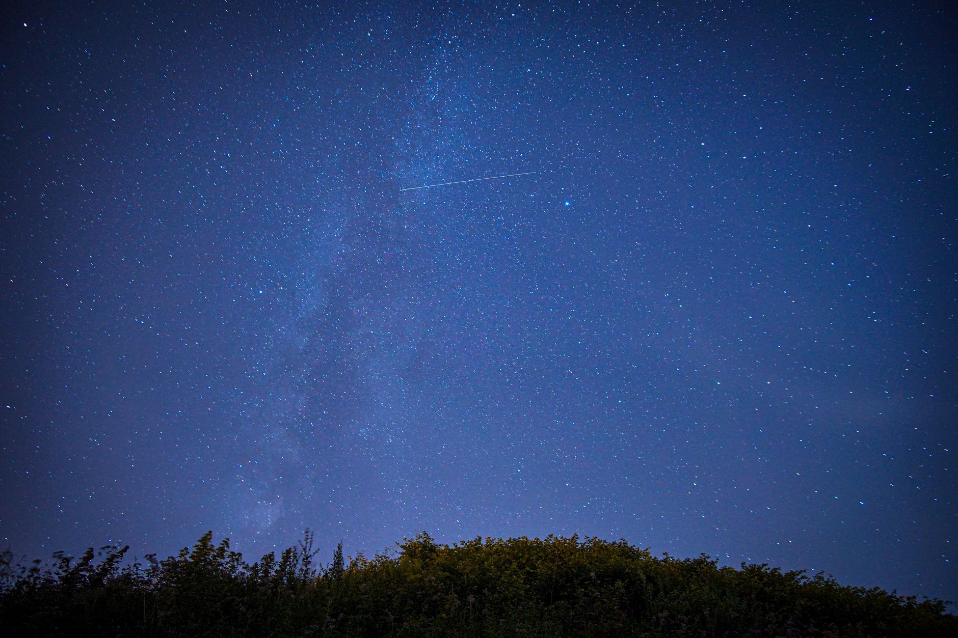 Beautiful starry night sky featuring a shooting star and the Milky Way over a silhouette of trees.