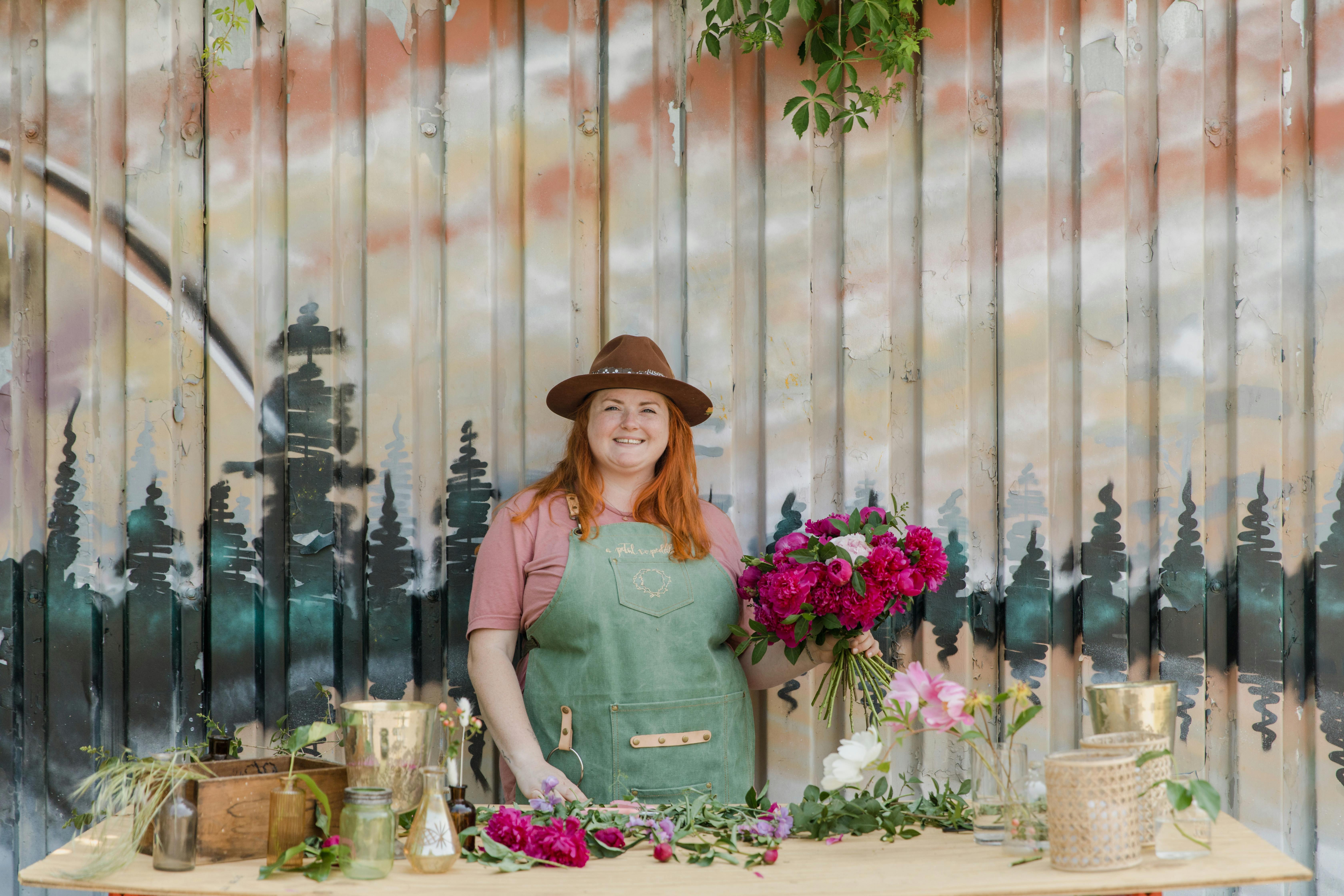 A smiling florist arranges vibrant flowers at an artistic outdoor workspace with a colorful backdrop.