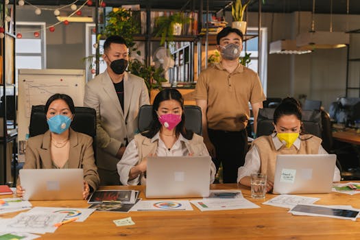 Diverse team working with laptops and papers, practicing safety by wearing face masks in an office setting.