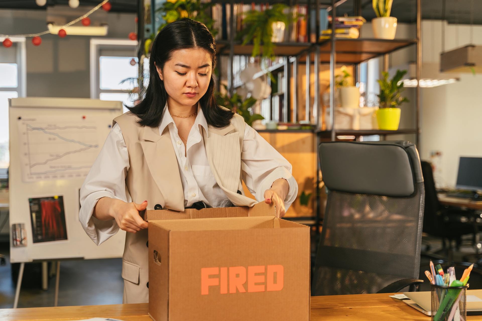 An Asian woman packs a box labeled 'FIRED', symbolizing job loss in an office setting.