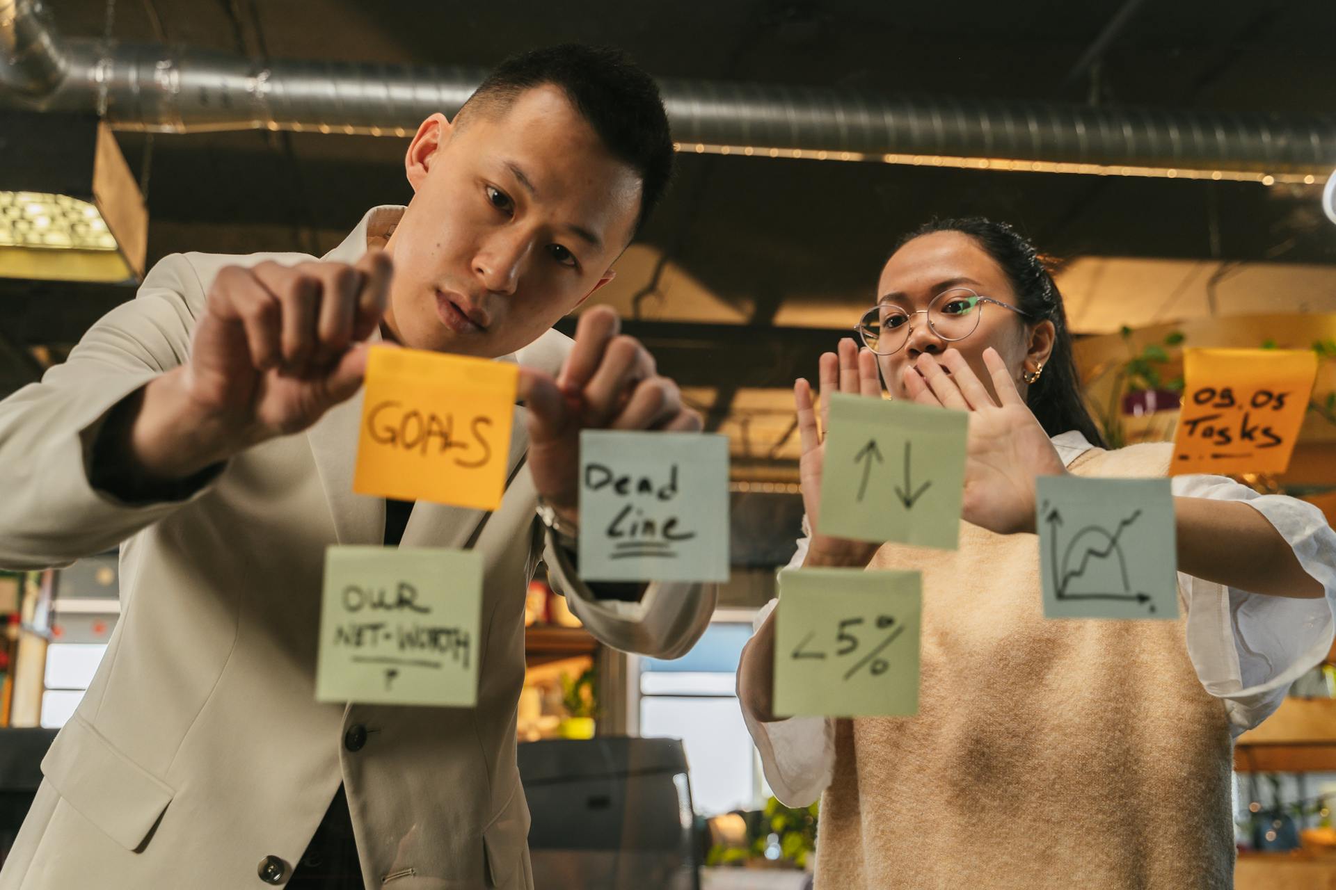 Two professionals organize tasks on glass using sticky notes in an office setting.