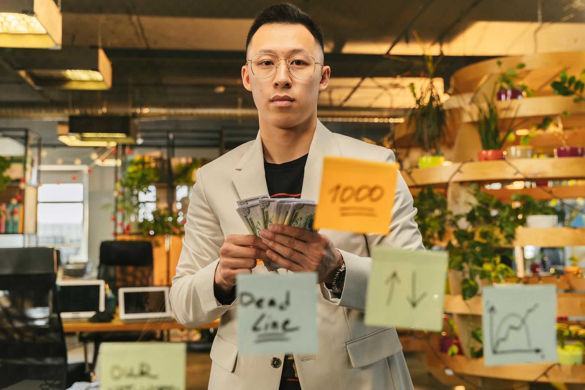 A businessman in a suit stands in a vibrant office, holding cash with sticky notes on glass.