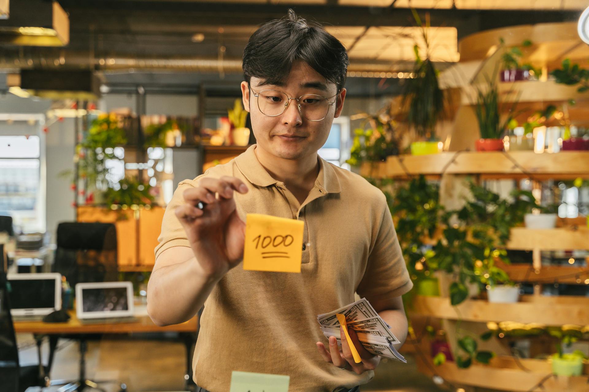 Businessman holds stack of cash and sticky note in a modern office setting.