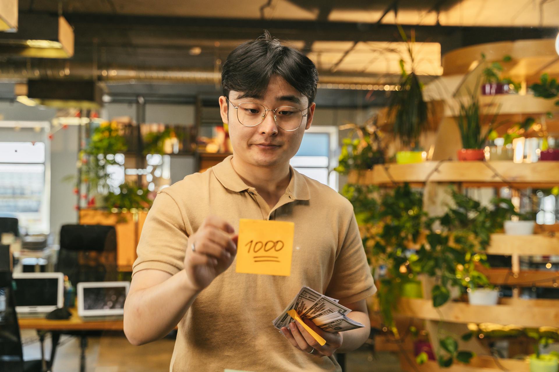 Asian man in a modern office holding cash and a sticky note with '1000', surrounded by plants.