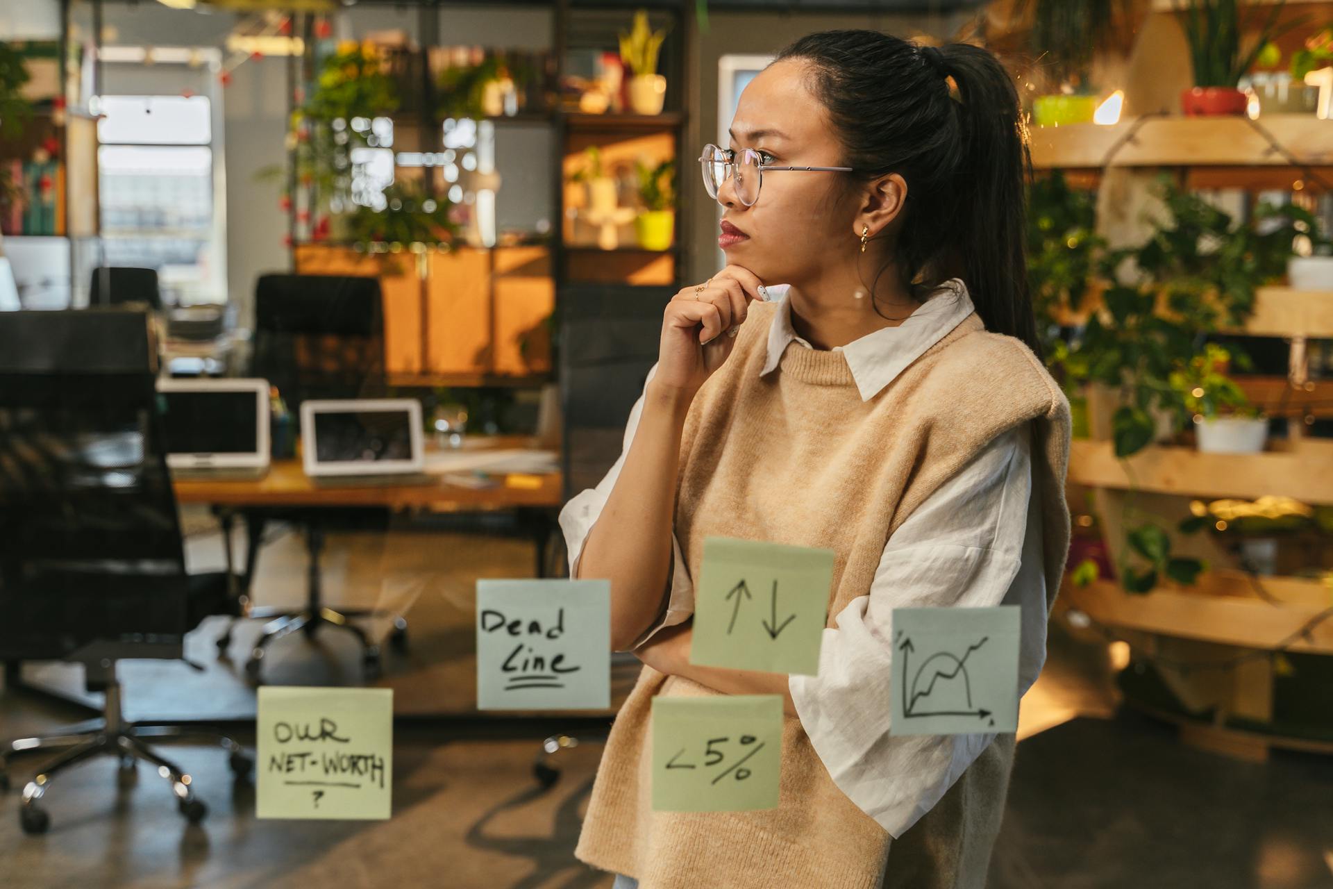 Asian woman in office examines sticky notes on glass, contemplating financial graphs.