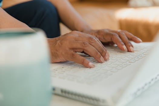 A focused shot of hands typing on a white laptop keyboard in a cozy indoor setting.