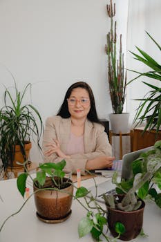 Asian woman with a smile sitting at a desk surrounded by plants, epitomizing a modern business setting.