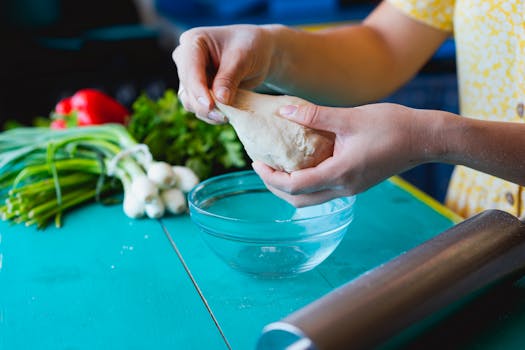 A person kneading dough in a colorful kitchen setup with fresh vegetables nearby.