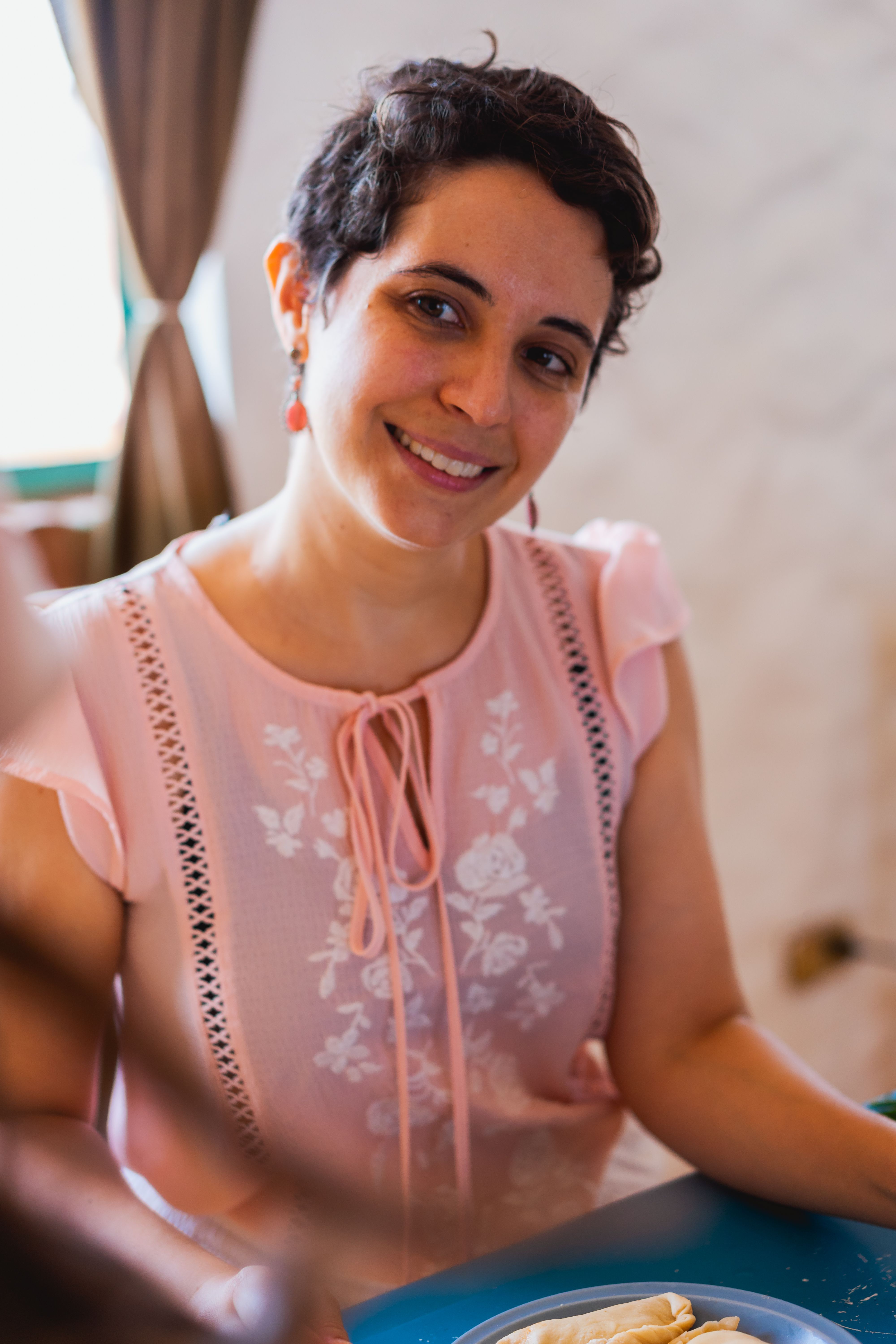A cheerful woman in a pink sleeveless top sitting at a table indoors, smiling.