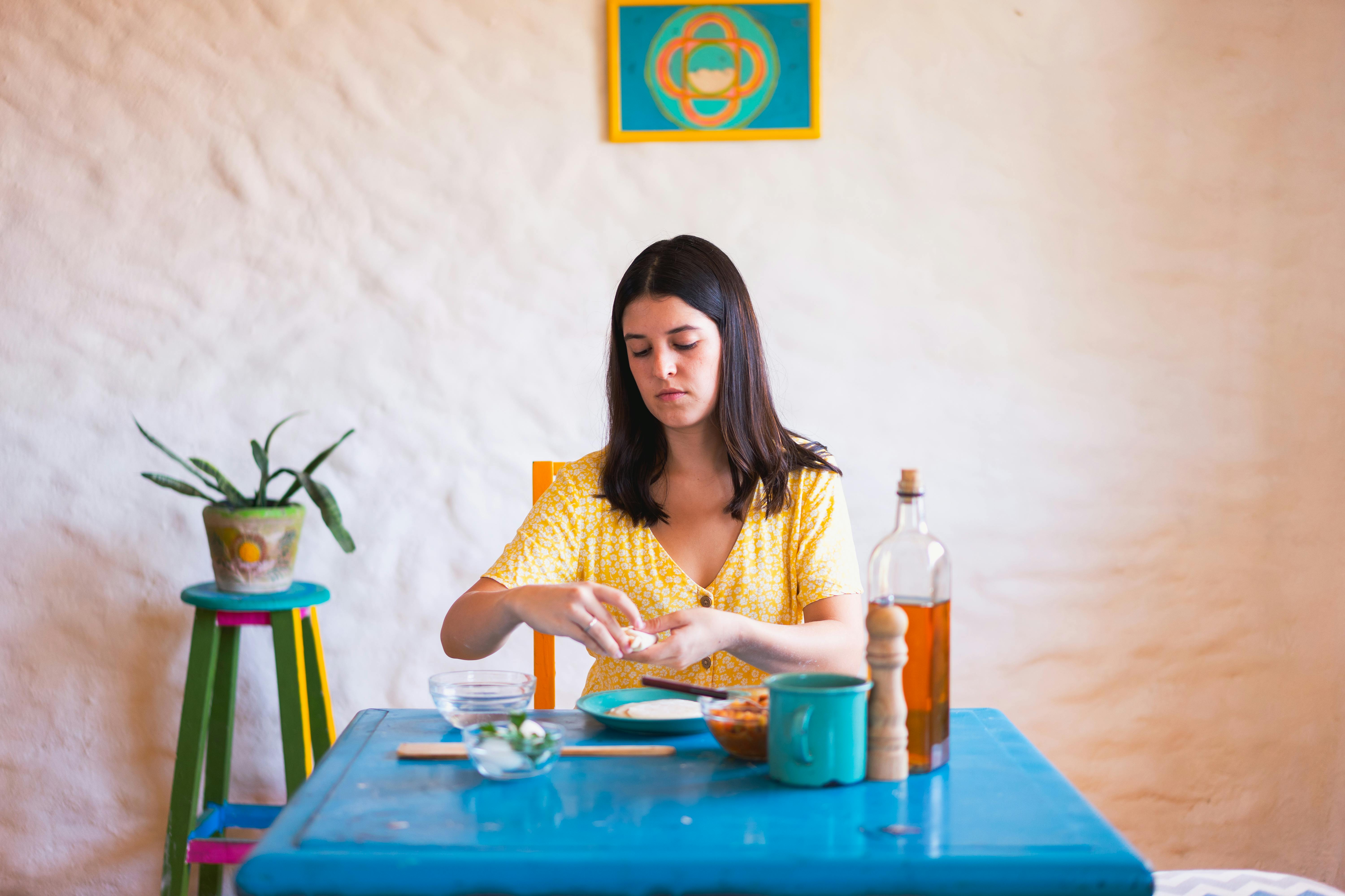 A young woman prepares traditional empanadas at a turquoise table indoors, surrounded by colorful decor.