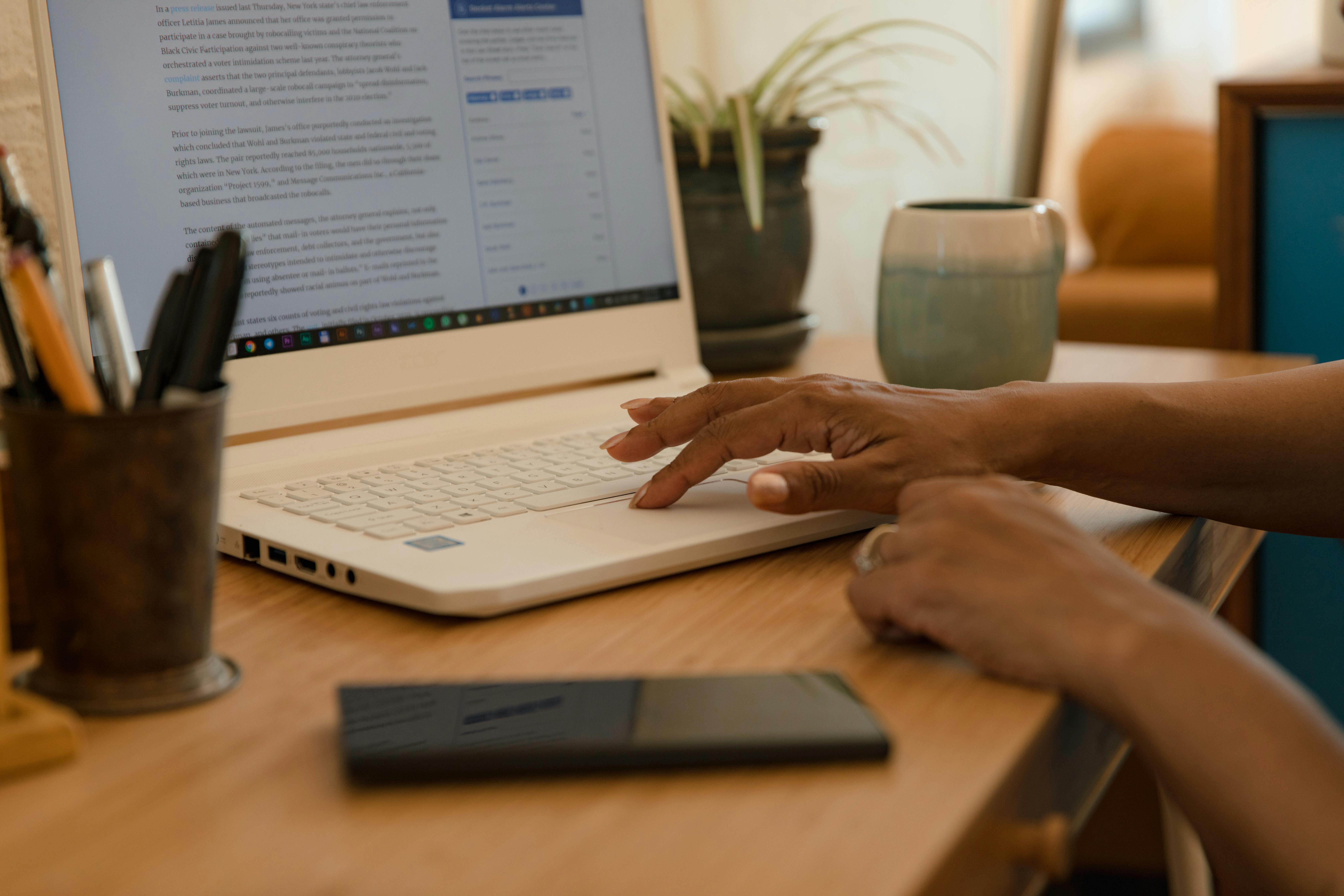 Hands typing on a laptop on a desk indoors with a mug and phone nearby.