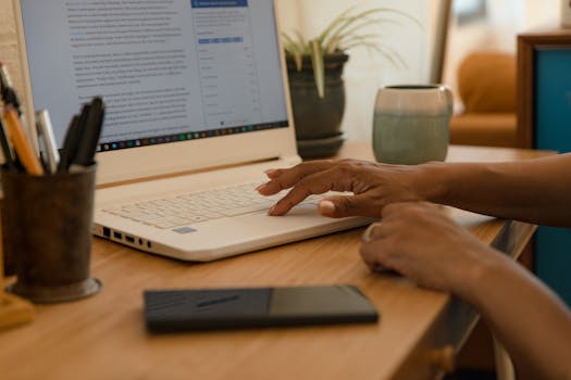 Hands typing on a laptop on a desk indoors with a mug and phone nearby.