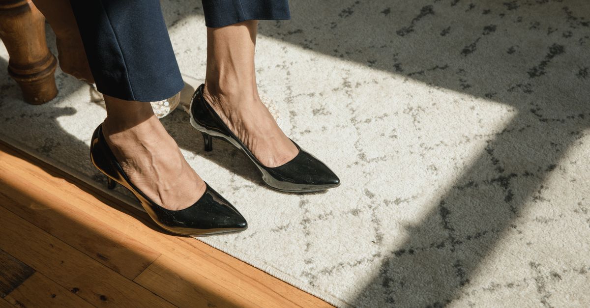 A close-up of feet in elegant black high heels standing on a patterned carpet in sunlight.