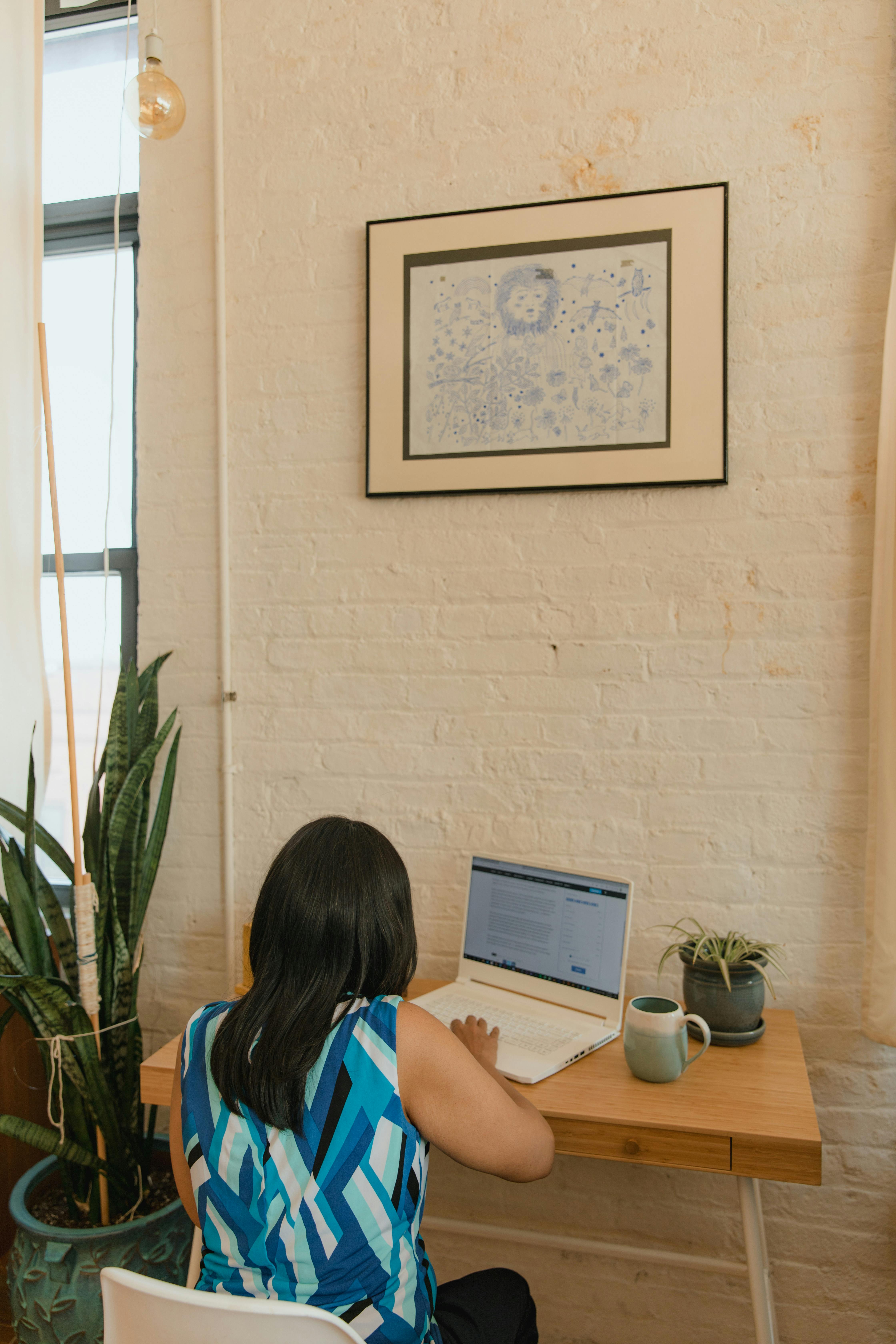 Back view of a woman in a blue blouse working at a laptop on a wooden desk in a cozy home office.