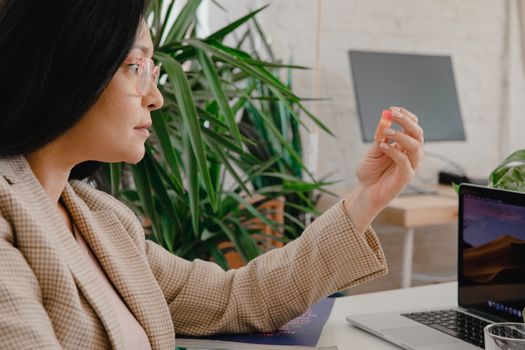 Asian woman in plaid blazer examines plastic item at modern workspace.