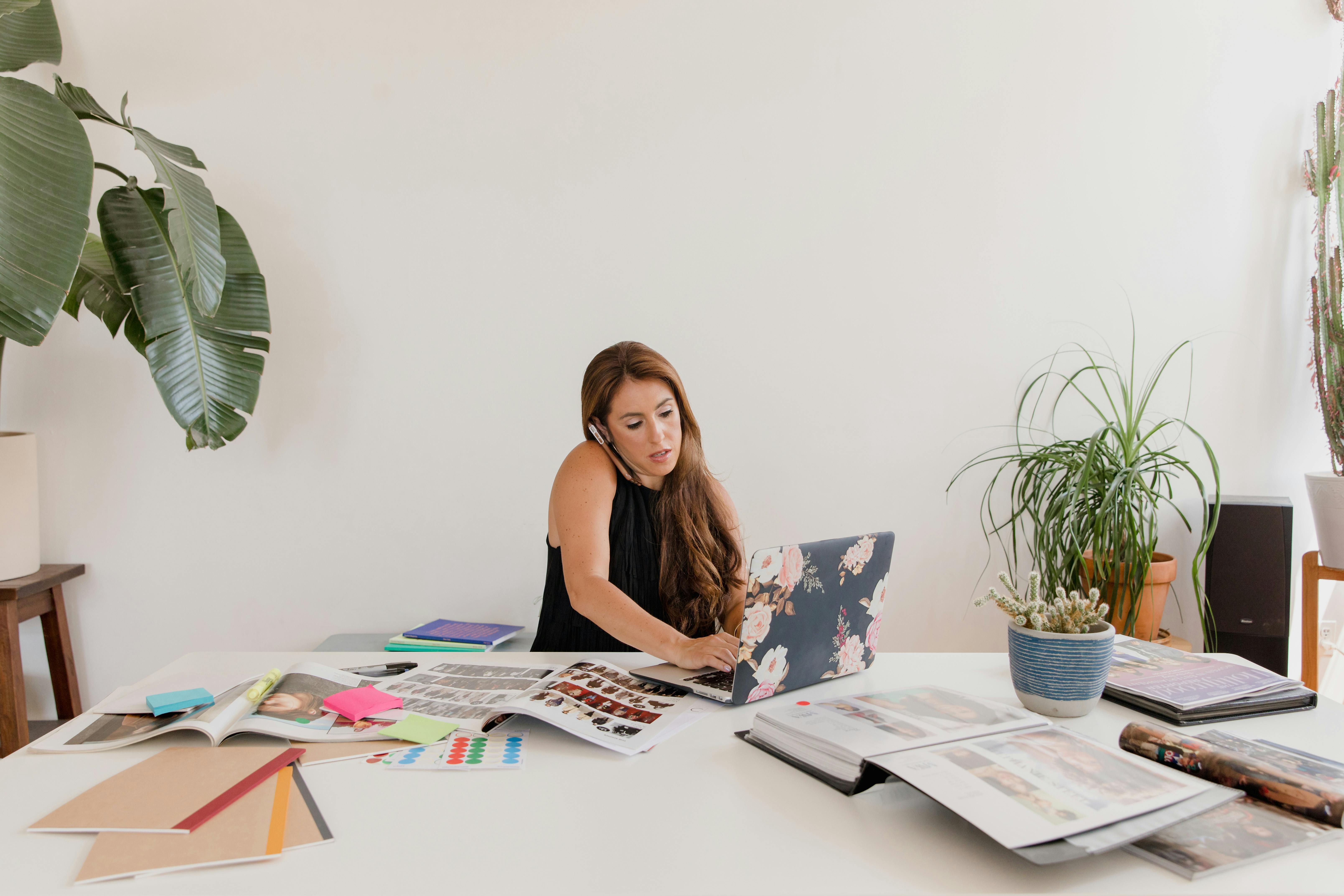 Woman working on a laptop while multitasking with calls, surrounded by creative materials and indoor plants in a home office.
