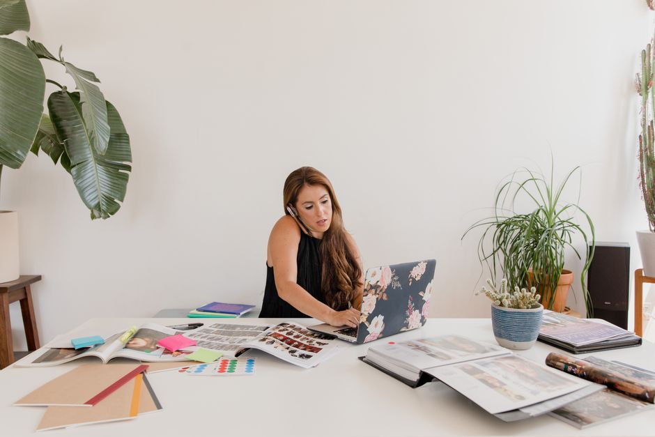 Woman working on a laptop while multitasking with calls, surrounded by creative materials and indoor plants in a home office. Woman working on a laptop while multitasking with calls, surrounded by creative materials and indoor plants in a home office.