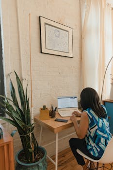 A woman focuses on her laptop in a cozy home office with a brick wall and potted plant.
