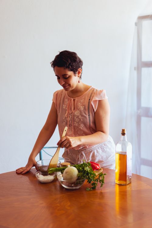 Free A woman in a peach blouse prepares a salad on a wooden table with fresh vegetables and cooking oil. Stock Photo