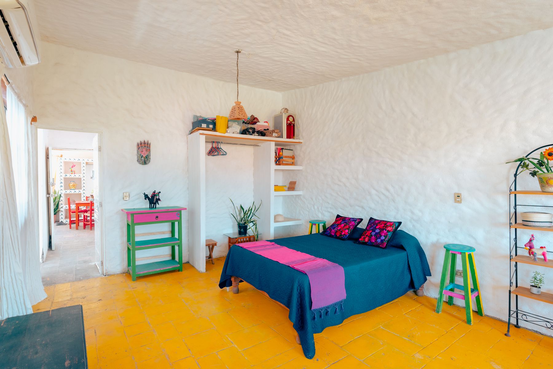 Bright maximalist bedroom with blue and pink bedding, yellow tile floor, colorful stools and black metal shelving