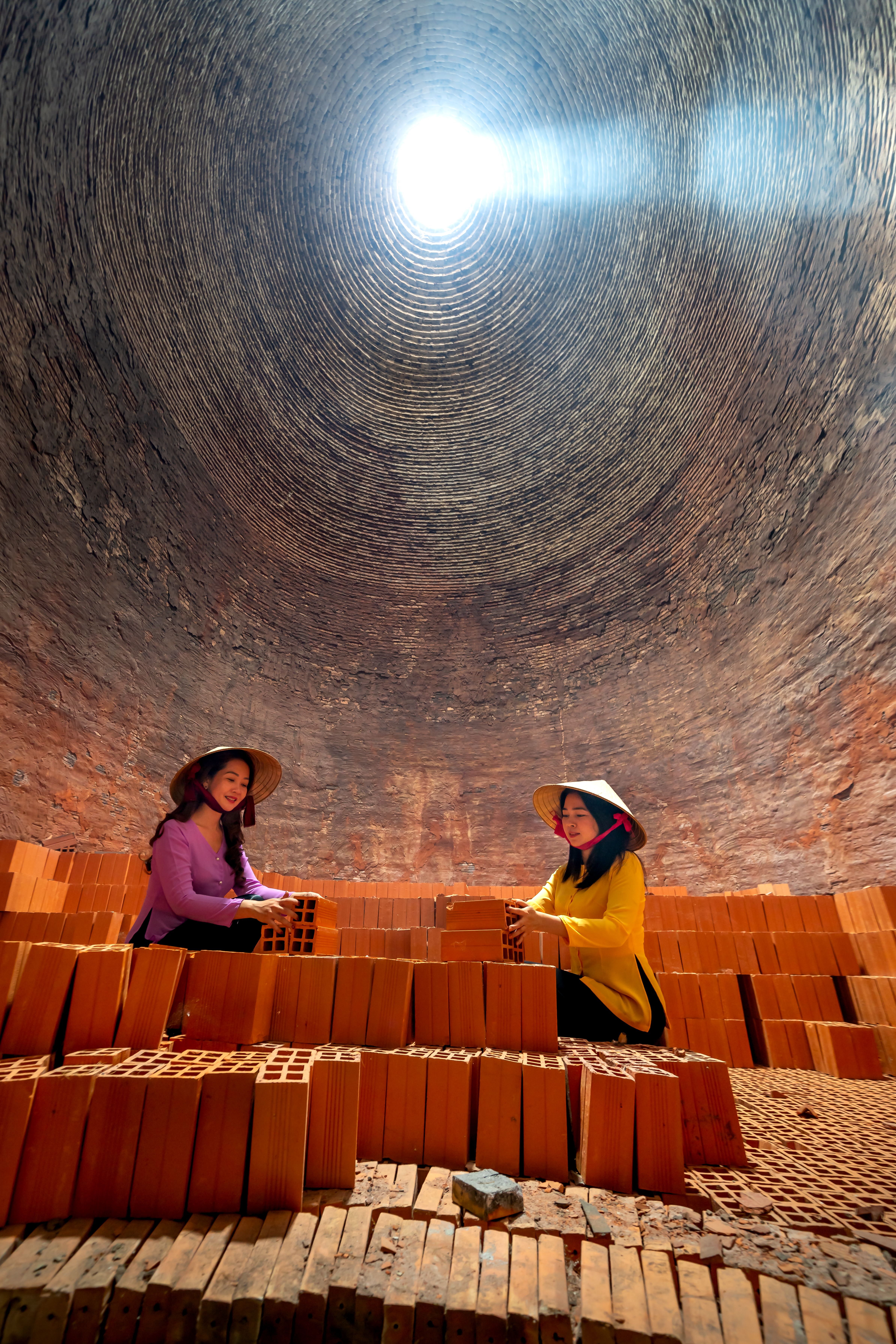 Two women in a brick kiln stacking bricks under a natural skylight.
