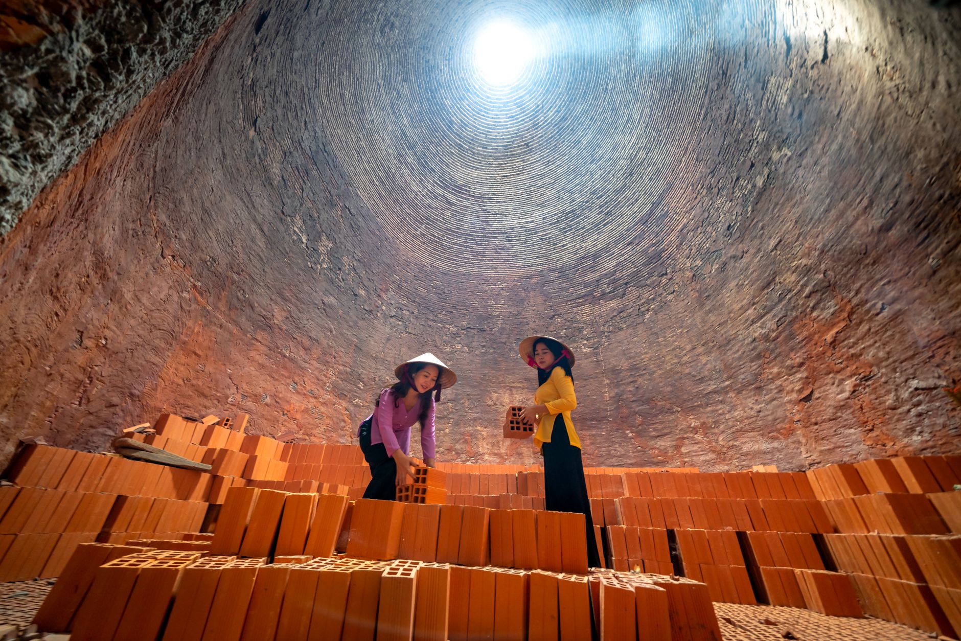 Two women stacking bricks inside a traditional Vietnamese brick kiln wearing conical hats.