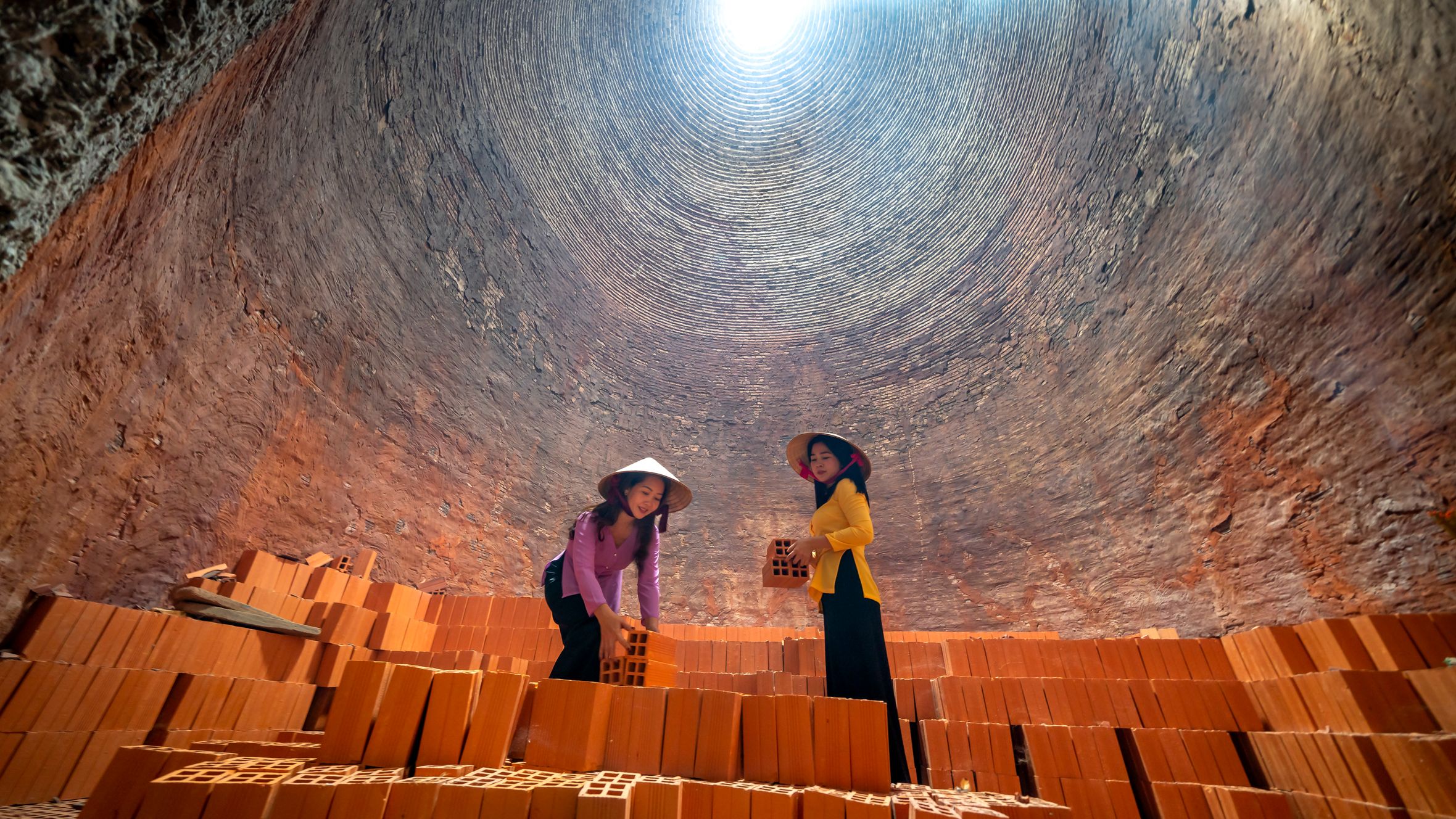 Two women stacking bricks inside a traditional Vietnamese brick kiln wearing conical hats.