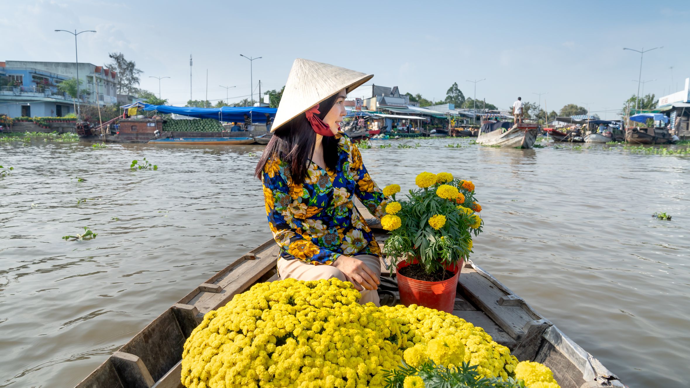 A woman wearing a conical hat selling vibrant yellow flowers on a boat in a busy river market.
