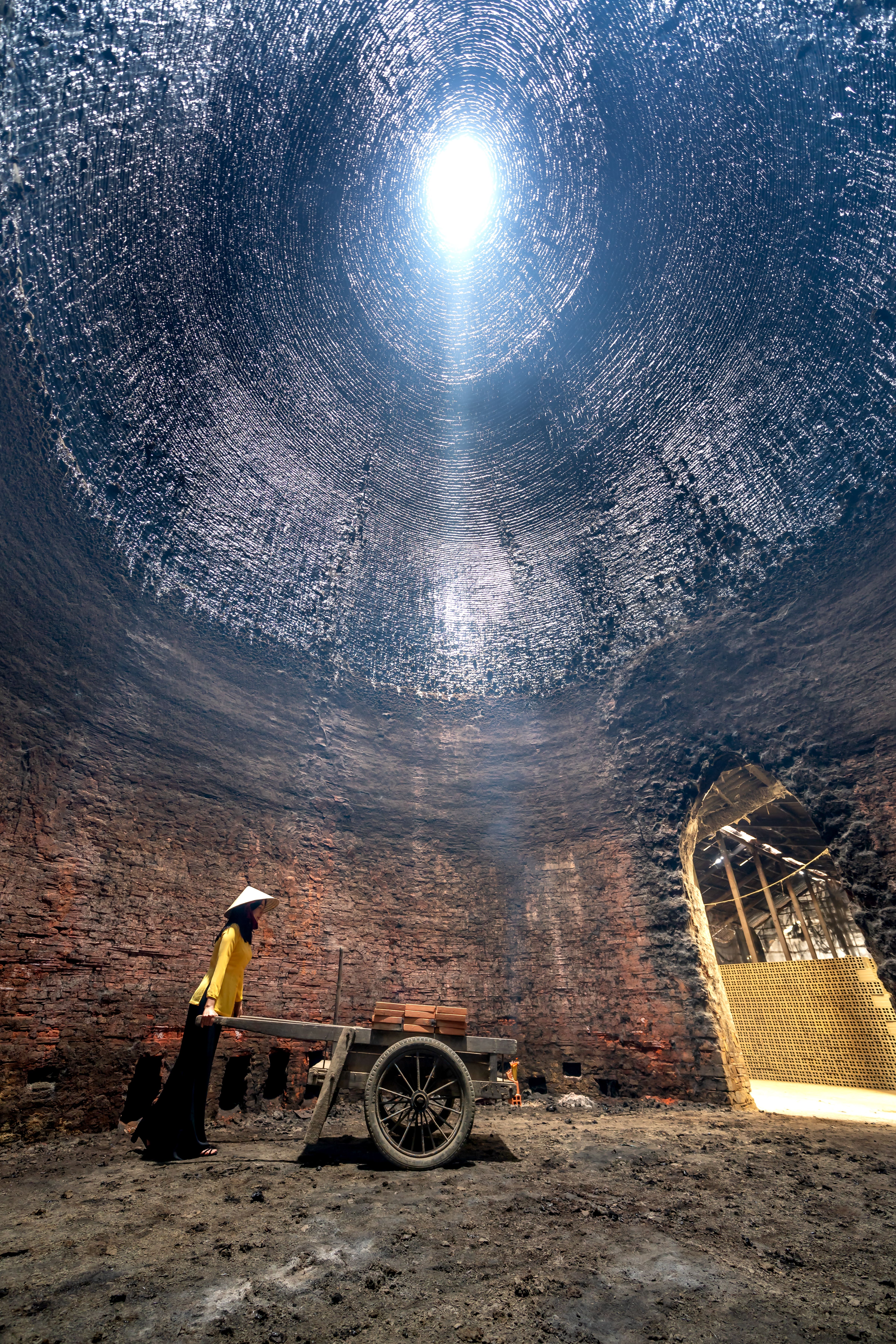 A woman pushing a wheelbarrow inside a traditional brick kiln in Vietnam, bathed in natural light.