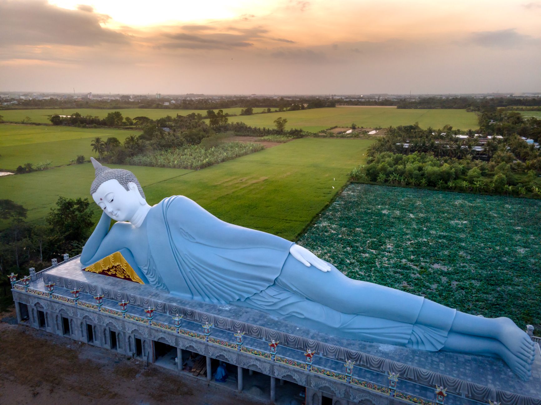 A serene aerial view of a giant reclining Buddha statue amid lush Vietnamese countryside at sunset.