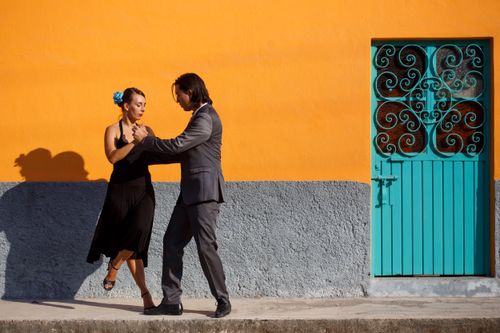 Free A couple performing a tango dance in front of a striking orange wall with a decorative turquoise door. Stock Photo