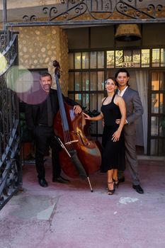 A stylish trio poses with a cello in an outdoor urban setting, showcasing elegance and music.