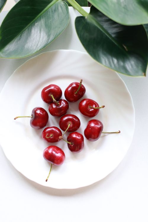 Free Juicy red cherries beautifully arranged on a simple white ceramic plate with lush green leaves. Stock Photo