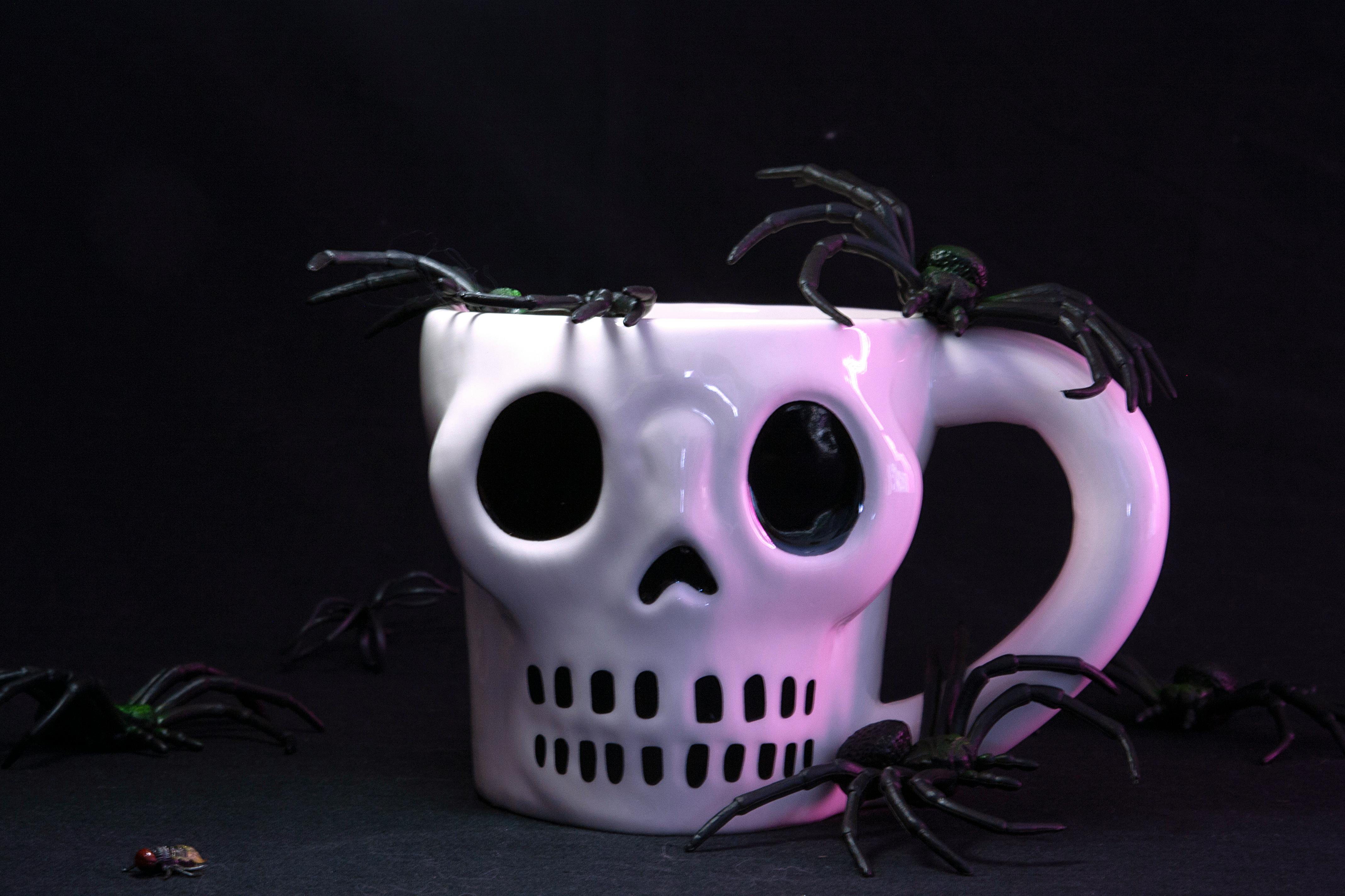 White skull-shaped mug surrounded by black spiders, creating a Halloween-themed scene.