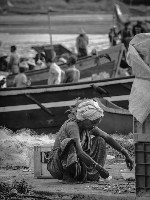 Free Black and white image featuring a woman crouching by boats at a bustling market. Stock Photo