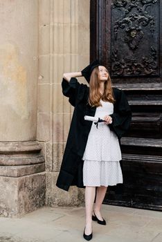 Confident graduate in cap and gown holding diploma outside historic building.