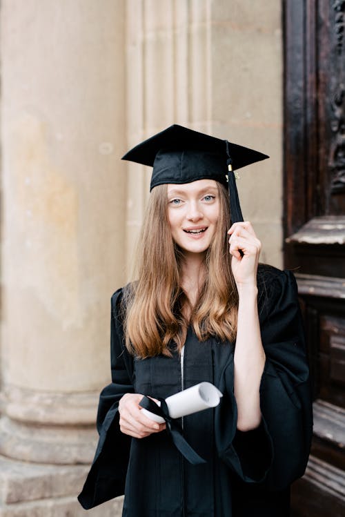 Free Woman Wearing a Black Academic Gown Holding Her Cap Stock Photo