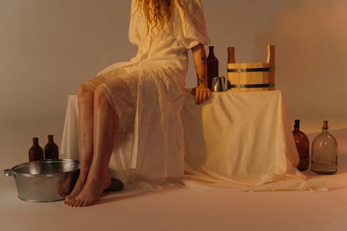 A Woman in White Lace Dress Sitting on a Chair Surrounded by Empty Glass Bottles