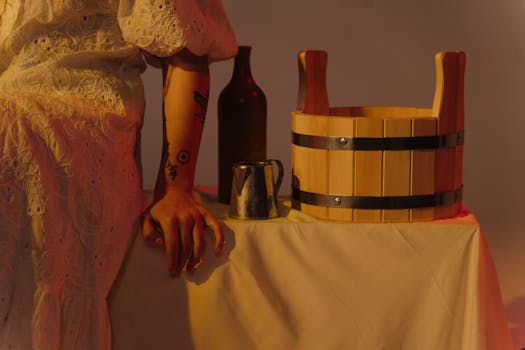 A person in vintage attire beside a wooden bucket, bottle, and jug on a table.