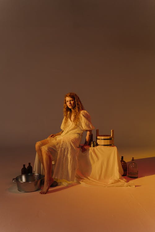 Woman in White Dress Sitting on a Chair Surrounded by Glass Bottles